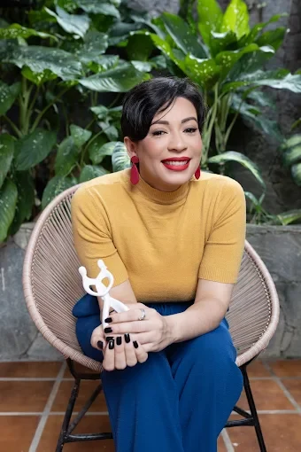 Laura Leite sorrindo, sentada em uma poltrona de vime entre plantas, segurando um pequeno objeto branco